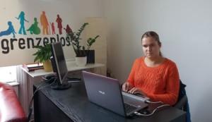 Kuvassa vapaaehtoistyöntekijä Susanna istuu pöydän ääressä, edessään kannettava tietokone. Huoneessa taustalla on Grenzenlosin logo ja huonekasvi.