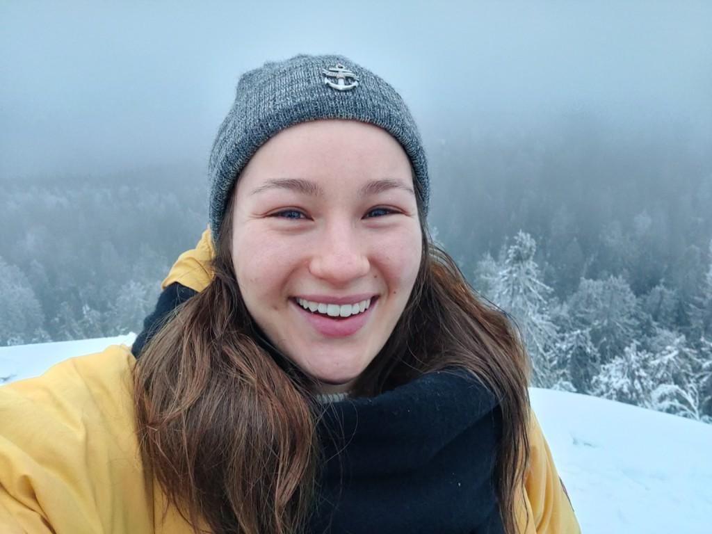 A smling person in a closeup photo, infront of a snowy forest scene.