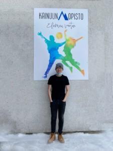 A person in front of a wall, in the wall there is a poster with text which says in Finnish "Kainuun opisto, for life".