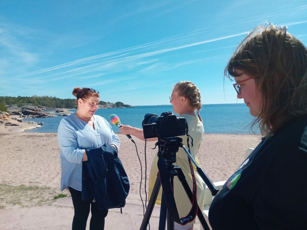 A person interviewing another person and a person standing behind a camera in the beach on a sunny day.