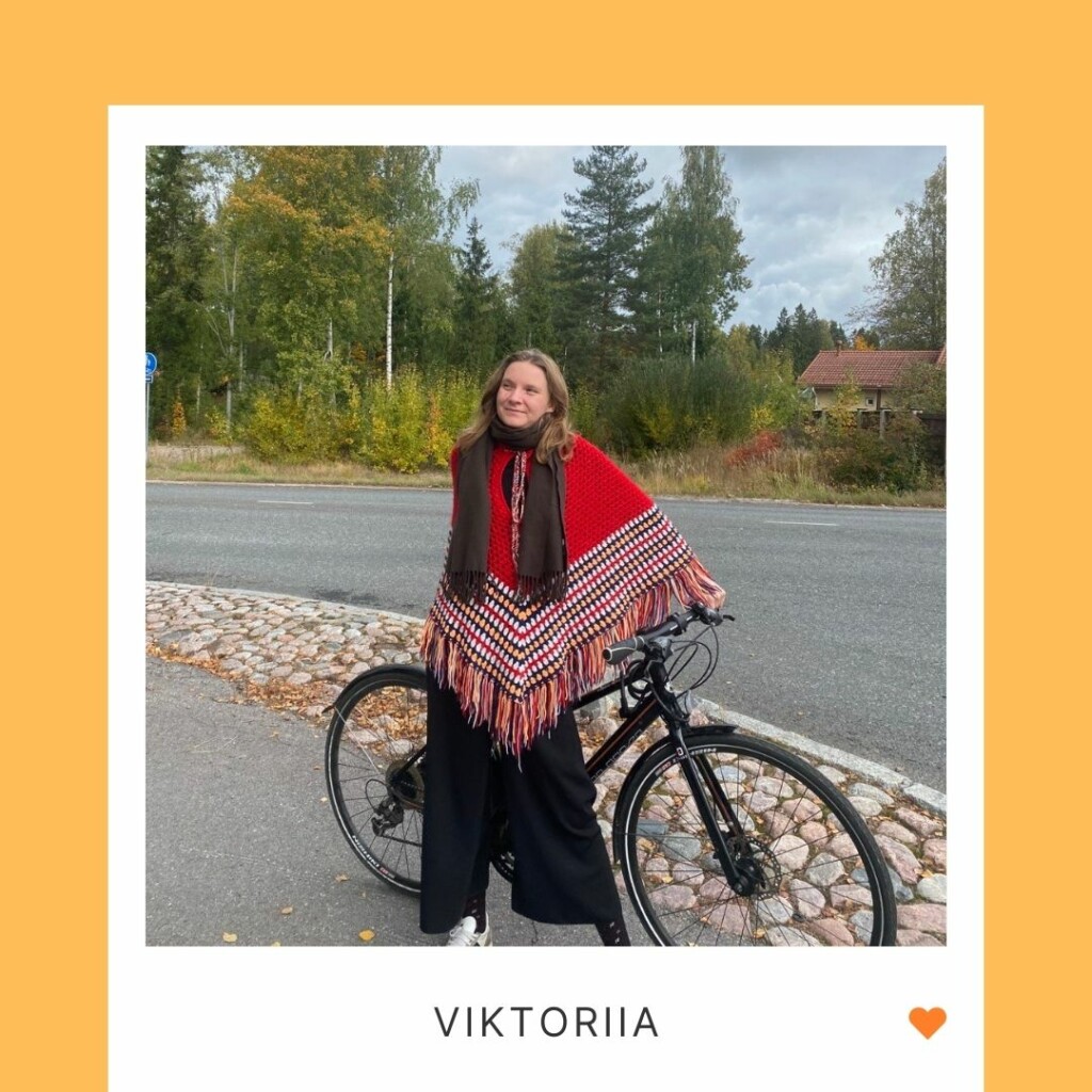 Hymyilevä henkilö seisoo kadulla polkupyörän edessä, alla teksti "Viktoriia" ja oranssi sydän.