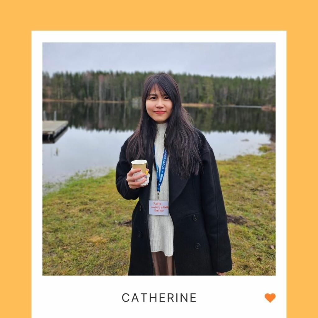 Hymyilevä henkilö seisoo ulkoalla, taustalla vettä ja metsää, kuvan alla teksti "Catherine" ja oranssi sydän. A smiling person standing outdoors, in the background there's water and forest, under the photo the text "Catherin" and an orange heart.