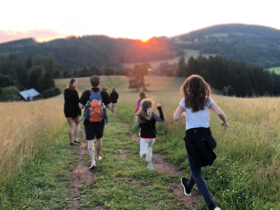 Children and adults moving away from the camera on a bath in the middle of a field during the sunset, in the background there are mountains.