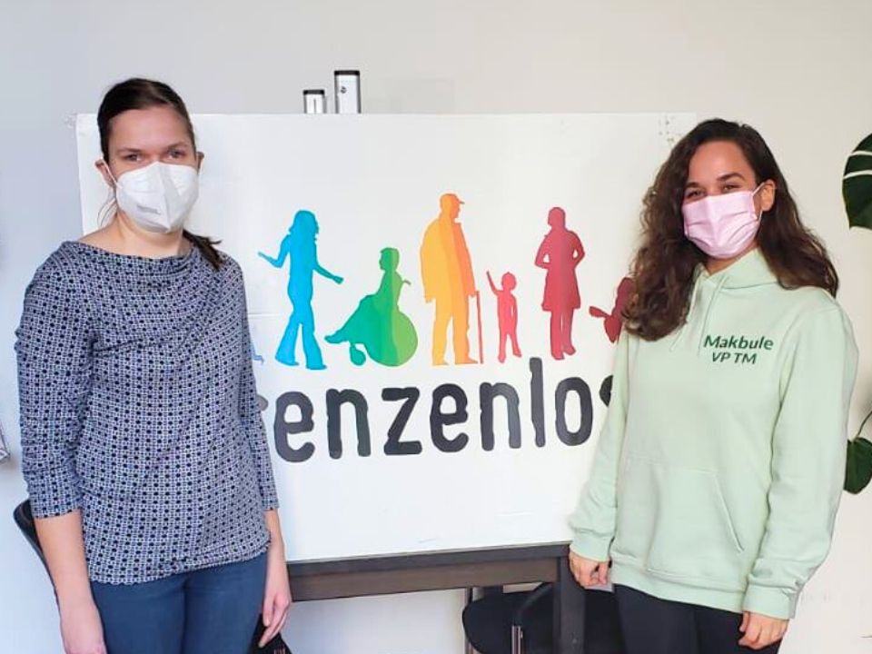 Two people standing in front of a sign that says has the logo of Grenzenlos on it, the people are looking at the camera and wearing face masks.