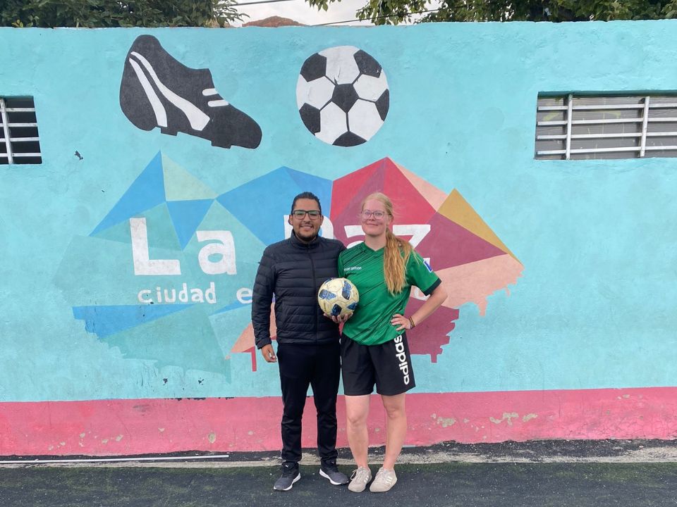 Two smiling people standing in front of a wall outdoors, one of them is holiding in thier hands a football.