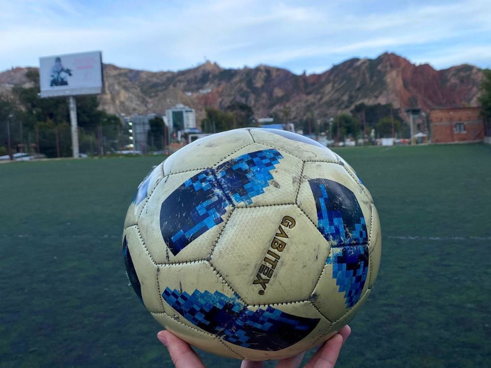 A hand holding a football. In the bacground, there is a football field and mountains.